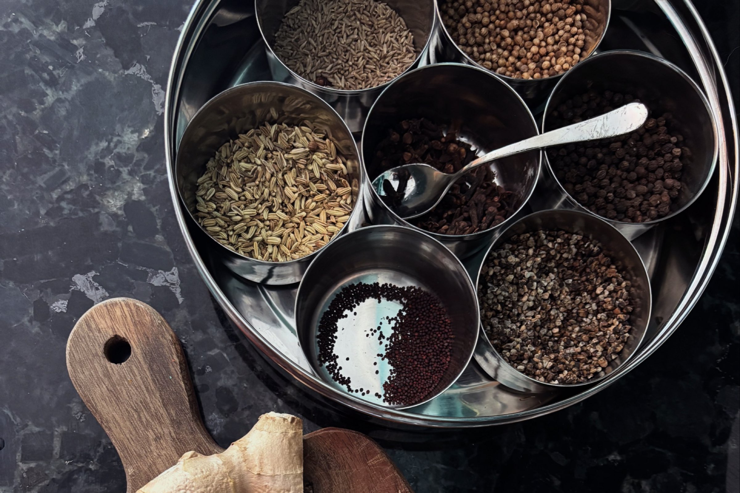 A masala dabba of whole spices with fresh ginger and turmeric on a wooden board
